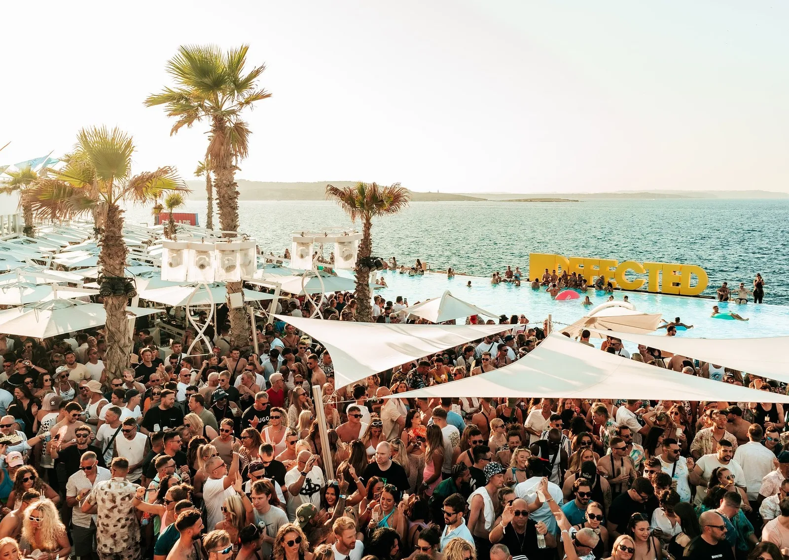 Crowd at a Defected Malta pool party by the sea, with palm trees and the Defected sign